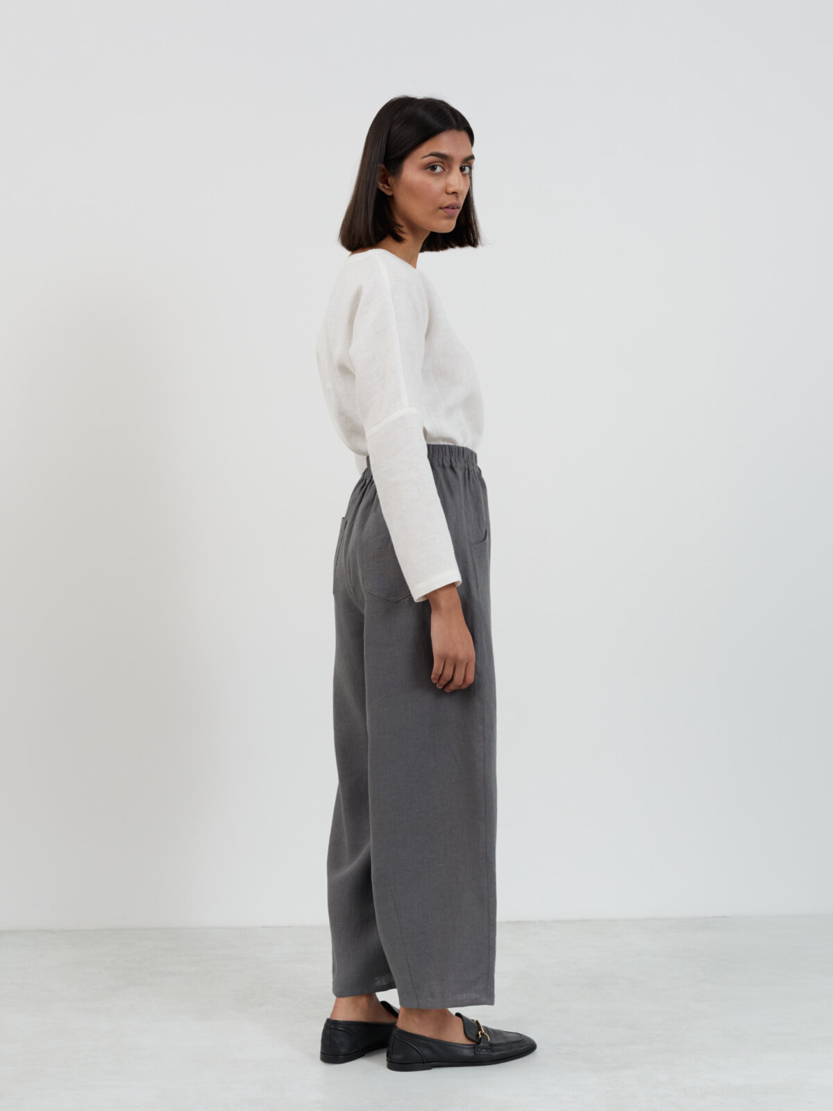 Three-quarter back view of Haven linen barrel leg pants in charcoal showing the side seam, back pocket placement, and ankle-length hem with black loafers
